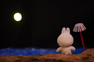 une poupée de lapin debout à côté d'un parapluie regardant la mer bleue la nuit avec la lumière de la lune. photo