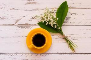 table basse du matin avec bouquet de fleurs de muguet. tasse jaune avec café noir décoré de fleurs printanières sur table en bois, vue de dessus. photo