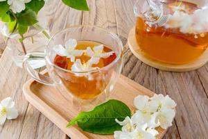 une tasse de thé au jasmin naturel dans une tasse en verre sur un plateau en bois avec des fleurs blanches. une table rustique avec une théière et des brins de jasmin dans un vase. photo