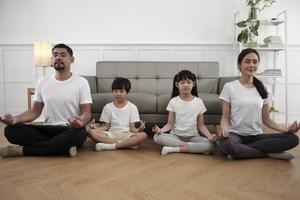 une famille thaïlandaise asiatique heureuse, des parents et des enfants s'assoient sur le sol du salon, ferment les yeux ensemble, méditent tranquillement en position du lotus et pratiquent un yoga sain, un week-end de style de vie à la maison. photo