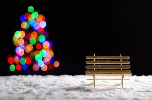 banc en bois lorsqu'il neige sur le sol en hiver et lumières bokeh colorées de l'arbre de noël. photo