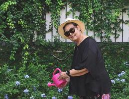 femme asiatique âgée heureuse et en bonne santé portant un chemisier noir, des lunettes de soleil et un chapeau, tenant un arrosoir rose arrosant des plantes dans le jardin, souriant et regardant la caméra. photo