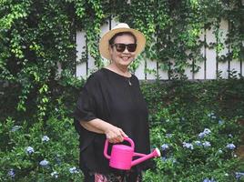 femme asiatique senior heureuse et en bonne santé portant un chemisier noir, des lunettes de soleil et un chapeau, tenant un arrosoir rose dans le jardin, souriant et regardant la caméra. photo