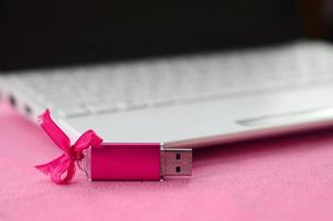 carte mémoire flash usb rose brillant avec un arc rose se trouve sur une couverture de tissu polaire rose clair doux et poilu à côté d'un ordinateur portable blanc. conception de cadeau féminin classique pour une carte mémoire photo