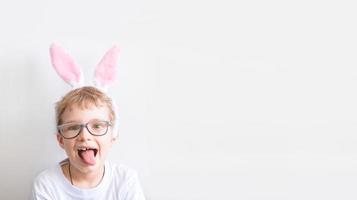 un garçon dans une tenue de lapin. un enfant heureux dans des verres dans un t-shirt blanc dans des oreilles de lapin montre la langue sur un fond blanc. concept de joyeuses pâques avec place pour le texte. photo