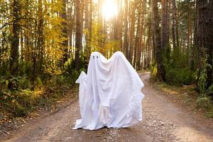 un enfant dans des draps avec une découpe pour les yeux comme un costume de fantôme dans une forêt d'automne fait peur et terrifie. un gentil petit fantôme rigolo. Fête d'Halloween photo