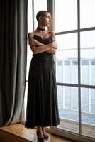 portrait d'une jeune femme triste aux cheveux courts debout regardant par la fenêtre dans une robe noire photo