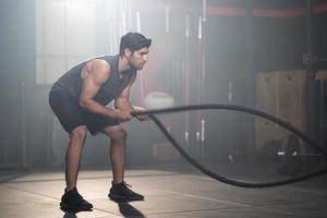 de beaux hommes caucasiens adultes balancent des cordes de combat pour faire de l'exercice crossfit à l'intérieur de la salle de fitness pour s'entraîner pour la force du corps et les muscles des bras fermes et une bonne santé physique du corps. photo