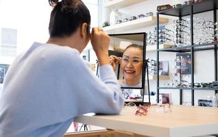 mise au point sélective au miroir. les femmes asiatiques âgées âgées sourient et regardent le miroir tout en choisissant de beaux essais de montures de lunettes à l'intérieur du magasin d'optique, du magasin. vue oculaire pour personnes âgées, soins de santé. photo