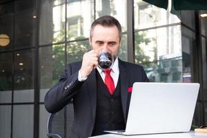 photo en plein air d'un homme d'affaires caucasien qui boit du café chaud le matin au café tout en utilisant un ordinateur portable pour travailler et consulter ses e-mails. routine de travail avec fond de zone d'affaires urbaine.