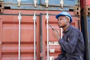 mise au point sélective sur le visage d'un travailleur logistique africain noir portant un équipement de sécurité, vérifier et inspecter l'état du conteneur tout en parlant et communiquer avec son équipe par appareil radio. photo