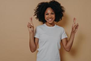 femme afro-américaine pleine d'espoir isolée sur fond de studio beige croisant les doigts faisant un vœu photo