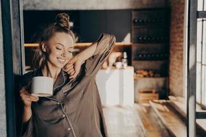 Heureuse belle femme brune qui s'étire avec une tasse de café à la main en se tenant debout dans la cuisine photo