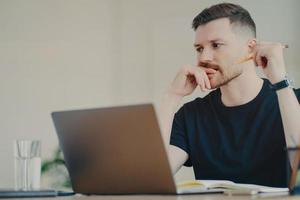 homme barbu sérieux étudiant aime étudier le processus a une expression réfléchie travaille en freelance vêtu d'un t-shirt noir décontracté écrit des notes utilise un ordinateur portable moderne pose dans un espace de coworking photo