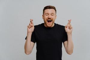 homme européen excité optimiste avec barbe assuré que le souhait se réalisera garde les doigts croisés attend des résultats positifs garde la bouche ouverte les yeux fermés anticipe le miracle isolé sur fond bleu photo