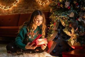 une adolescente heureuse ouvre une boîte-cadeau rouge assise sous un sapin de noël à minuit photo