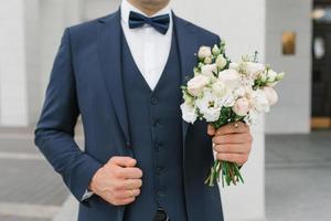 le marié en costume bleu tient dans sa main un bouquet de mariage délicat pour sa mariée bien-aimée photo