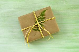 coffret cadeau enveloppé de papier kraft et ruban blanc et jaune avec feuille de fougère sur fond de bois vert, vue de dessus. photo