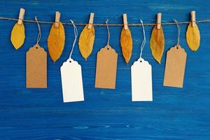 cinq étiquettes ou étiquettes de prix en papier vierge marron et blanc accrochées à une corde avec des feuilles d'automne jaunes sèches sur le fond en bois bleu. photo