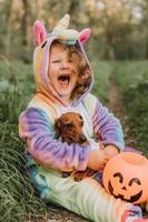 petite fille dans un costume d'halloween de licorne arc-en-ciel et un teckel dans une robe avec un panier de citrouille pour les bonbons sont assis sur une souche au coucher du soleil de la forêt. fabuleuse forêt magique merveilleuse. espace pour le texte photo