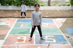 jolie petite fille asiatique jouant à la marelle en plein air. jeu d'activité amusant pour les enfants sur l'aire de jeux à l'extérieur. sport de rue d'été pour les enfants. mode de vie d'enfance heureux. photo