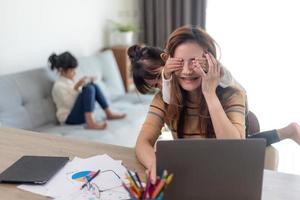 une femme est assise à la table du bureau à domicile pendant le verrouillage, travaillant sur un ordinateur portable. un enfant enjoué distrait du travail, couvrant les yeux de sa mère, un enfant faisant du bruit et demandant l'attention d'une mère occupée photo