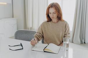 Une jeune femme rousse sérieuse prend des notes dans le bloc-notes est assise à table photo