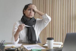 jeune femme européenne malsaine employée de bureau tenant un thermomètre numérique et touchant le front photo