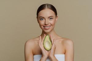 concept de beauté et de soins personnels. une femme européenne calme et souriante aux cheveux peignés tient un avocat comme recommandation pour un masque facial nourrissant enveloppé dans une serviette de bain isolée sur fond de studio beige photo