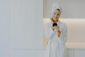 une jolie femme applique une crème hydratante sur le visage prend soin de son teint et de sa peau tient un miroir porte un peignoir blanc sourit doucement regarde pensivement pose sur des meubles modernes à la maison photo