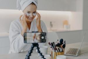 photo d'une blogueuse influenceuse enregistre une vidéo comment appliquer des tampons de beauté sous les yeux regarde la caméra d'un smartphone enseigne aux adeptes la procédure de soins de la peau pose contre l'intérieur de la maison en peignoir