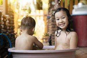 la sœur aînée et le frère prenaient joyeusement un bain en plastique le matin. prendre une douche dehors par une chaude journée. frères et sœurs sont heureux de jouer dans l'eau. enfants s'amusant à jouer dans l'eau photo
