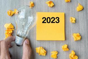 2023 mots sur une note jaune et du papier émietté avec un homme d'affaires tenant une ampoule sur fond de table en bois. nouvel an nouvelle idée créative, innovation, imagination, résolution et concept d'objectif photo