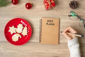 main de femme écrivant des plans de 2023 sur un ordinateur portable avec des biscuits de noël sur la table. noël, bonne année, objectifs, résolution, liste de choses à faire et concept de stratégie photo