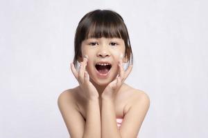 belle fille de gym asiatique riant en appliquant de la crème pour le visage avec une peau fraîche et propre sur fond blanc. soins du visage et spas, soins du visage, visage de fille asiatique photo