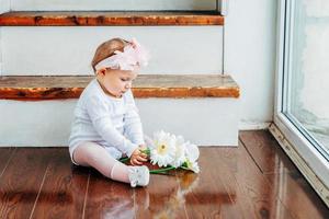 petite fille souriante d'un an portant une couronne de printemps assise sur le sol dans un salon lumineux près de la fenêtre et jouant avec des fleurs de gerbera. enfant heureux jouant à la maison. notion d'enfance. photo