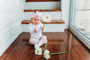 petite fille souriante d'un an portant une couronne de printemps assise sur le sol dans un salon lumineux près de la fenêtre et jouant avec des fleurs de gerbera. enfant heureux jouant à la maison. notion d'enfance. photo