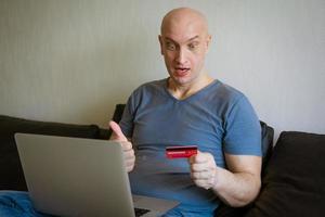 homme chauve émotionnel sur canapé avec ordinateur portable et carte de crédit en main photo