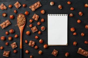 chocolat aux noisettes, un cahier avec des pages blanches et une cuillère en bois avec du cacao sur fond sombre, entouré de noix dans la coque et pelées. photo