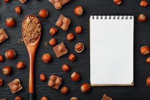 chocolat aux noisettes, un cahier avec des pages blanches et une cuillère en bois avec du cacao sur fond sombre, entouré de noix dans la coque et pelées. photo