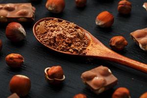 chocolat aux noisettes et une cuillère en bois avec du cacao sur fond sombre, entouré de noix dans la coque et pelées. photo