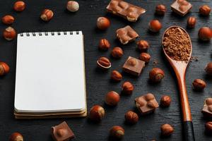 chocolat aux noisettes, un cahier avec des pages blanches et une cuillère en bois avec du cacao sur fond sombre, entouré de noix dans la coque et pelées. photo