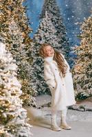 jolie fille blonde caucasienne en manteau de fourrure écologique blanc marchant dans la forêt de noël d'hiver avec des lumières. nouvel an, concept de conte de fées photo