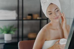 belle femme en bonne santé applique une crème hydratante, enveloppée dans une serviette de bain, regarde attentivement la réflexion dans le miroir, bénéficie d'un traitement de soins de la peau après la douche, pose dans la salle de bain. notion de beauté. photo