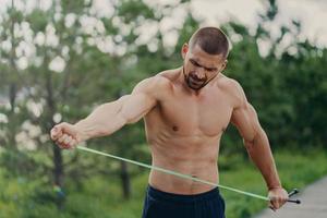 jeune bel homme européen fort pose au stade du parc, étire la corde à sauter, a le torse nu et les bras musclés, va faire des exercices physiques, pose en plein air. concept d'entraînement de rue photo