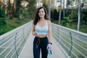 photo extérieure d'une femme mince en vêtements de sport fait du jogging près de la forêt, aime écouter de la musique dans les écouteurs de sa liste de lecture préférée, tient une bouteille d'eau et pose sur le pont. concept de style de vie sportif