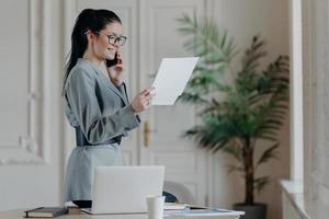 photo à l'intérieur d'une femme souriante et heureuse qui vérifie les informations des papiers porte un costume formel gris, des lunettes optiques concentrées sur le travail de recherche a une conversation téléphonique, se tient dans un espace de coworking