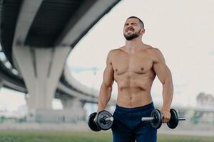 concept d'endurance et de puissance. un européen barbu motivé lève des haltères met tout en œuvre pour soulever des poids lourds serre les dents pose avec un équipement de sport sous le pont, a un corps musclé photo