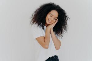 charmante belle femme au sourire tendre, s'appuie sur les paumes pressées, rêve de dormir, incline la tête, porte un t-shirt blanc, a une beauté naturelle, des dents parfaites, pose à l'intérieur. jeune femme afro rêveuse photo