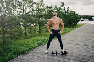 un homme musclé en bonne santé se tient le torse nu, lève la tête au-dessus et respire profondément, porte des vêtements de sport, s'entraîne avec des haltères, pose à l'extérieur. musculation, mode de vie actif, concept de remise en forme photo